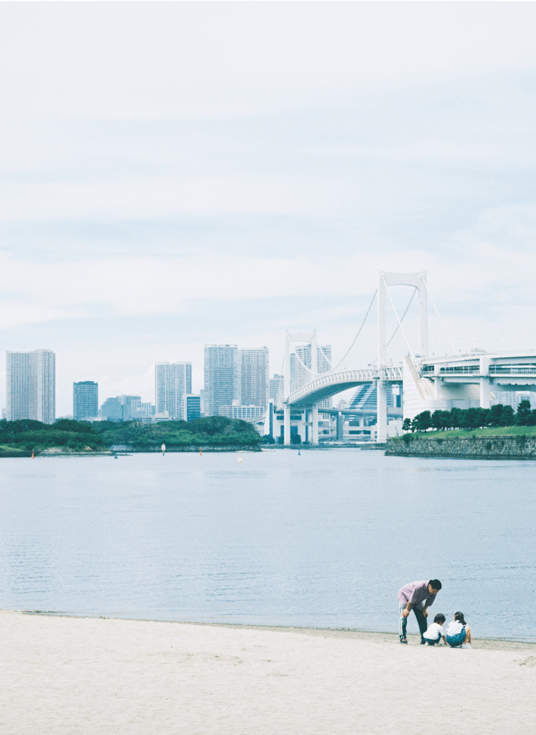 大人1人と子供2人が砂浜で遊んでいる背景には大きな橋と複数ビルが写っている様子。